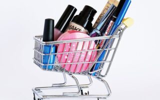 Miniature shopping cart filled with a variety of cosmetics on a white background.