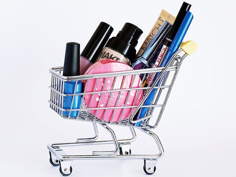 Miniature shopping cart filled with a variety of cosmetics on a white background.